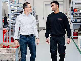 Zwei Mitarbeiter gehen durch eine Produktionshalle und lächeln sich an