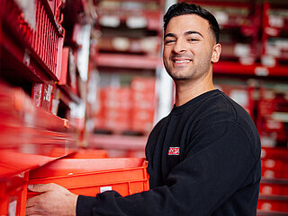 Mitarbeiter lächelt in die Kamera und schiebt rote Kiste in Regal