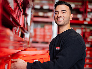 Mitarbeiter lächelt in die Kamera und schiebt rote Kiste in Regal