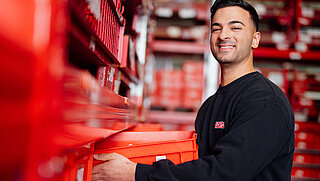 Mitarbeiter lächelt in die Kamera und schiebt rote Kiste in Regal