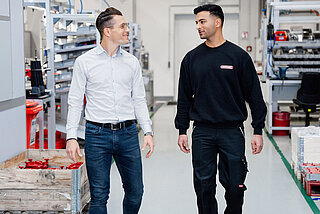 Zwei Mitarbeiter gehen durch eine Produktionshalle und lächeln sich an