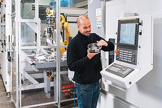 Mitarbeiter vor automatisierter Drehmaschine mit Drehteil in der Hand